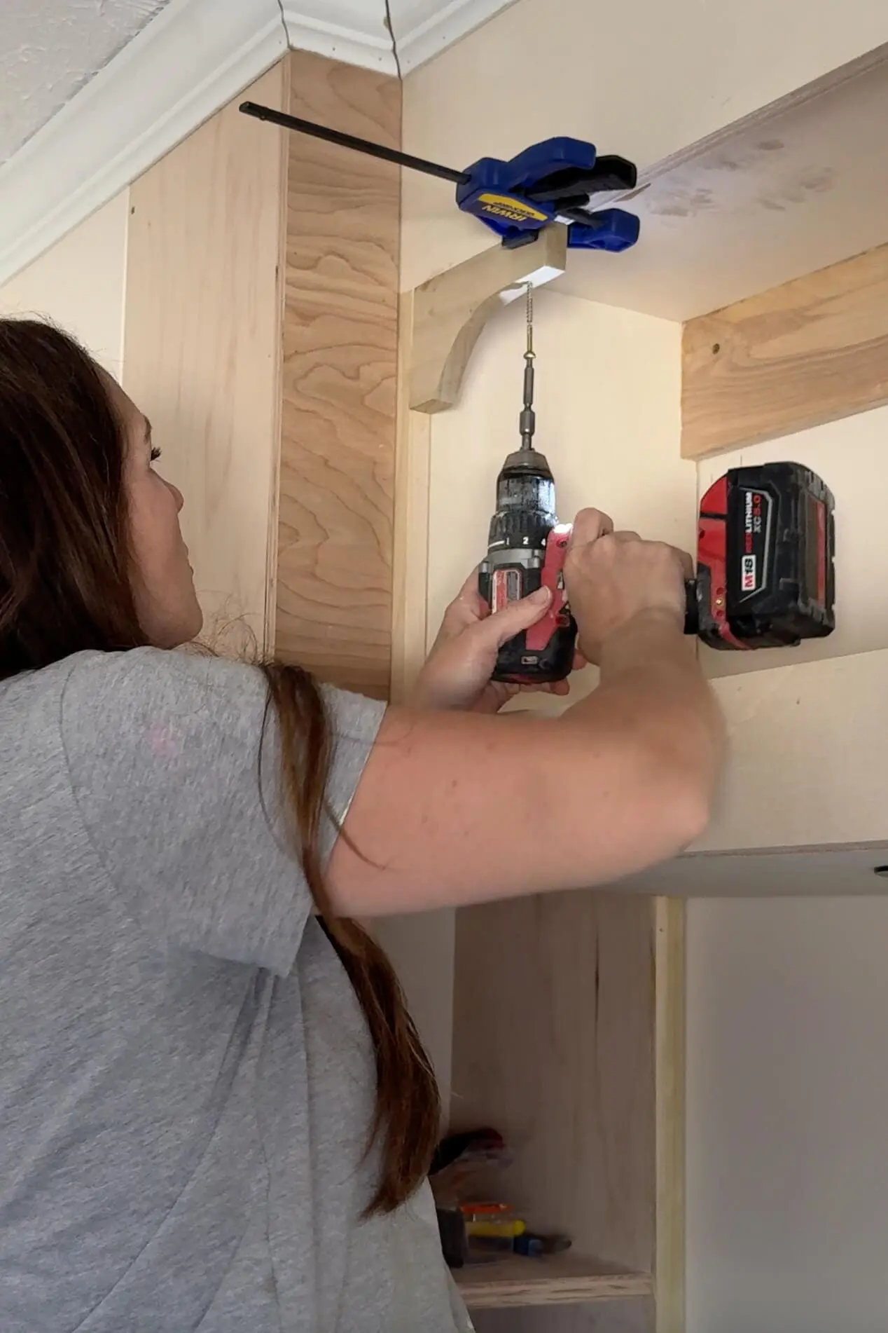 Securing a support bracket into the side of a built-in headboard with storage using a drill and clamp.